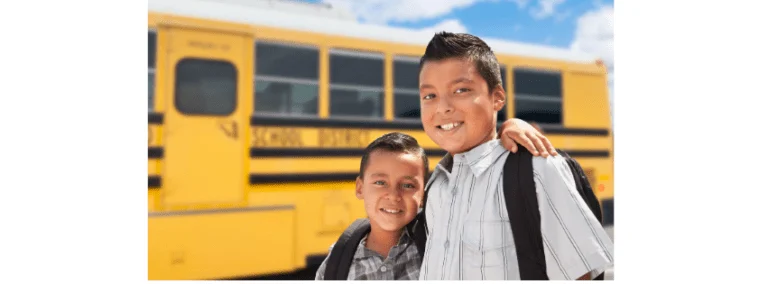 Two smiling Hispanic boys wearing backpacks in front of a yellow school bus