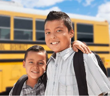 Two smiling Hispanic boys wearing backpacks in front of a yellow school bus