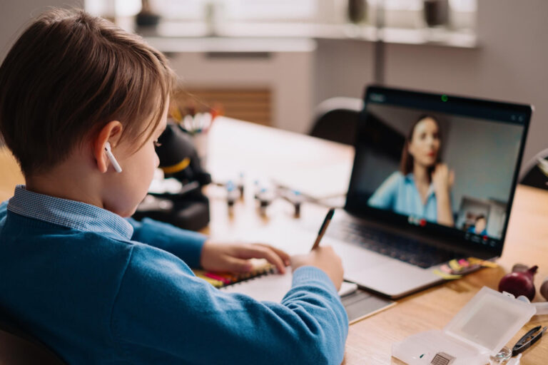 A child at a laptop in a home setting wearing a headset, with a tutor visible on the screen.